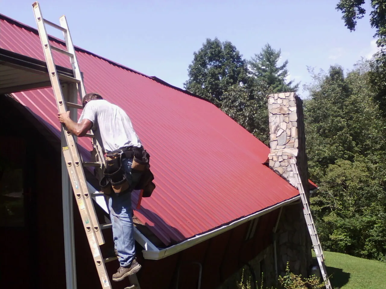 Roofer on a ladder beside a red metal roof and stone chimney — Soffit & Fascia Repair by Roofing Brothers in Rosendale